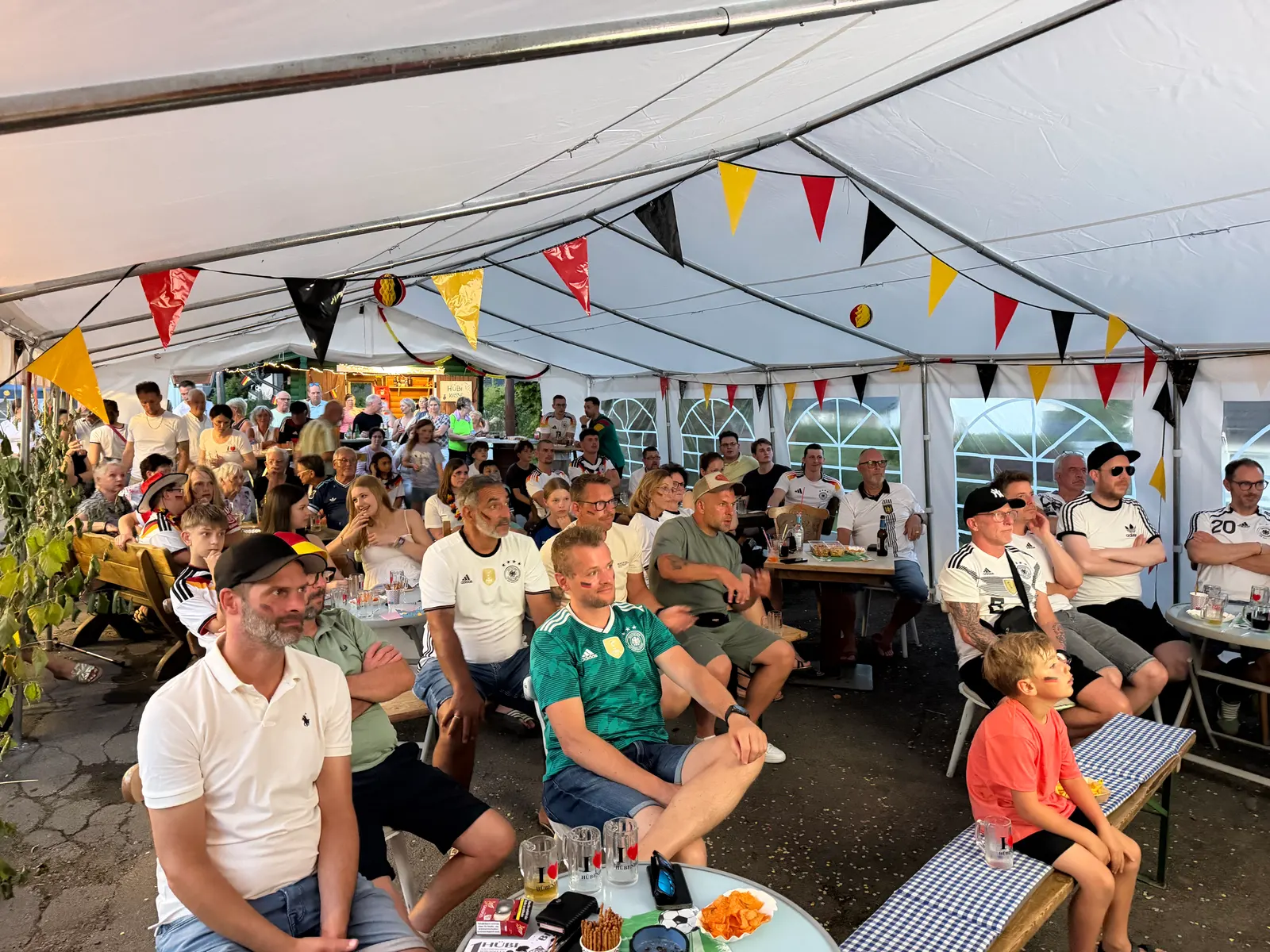 Volles Festzelt am Dorfplatz bei der WM-Kneipe: viele Menschen in Deutschland-Trikots verfolgen gemeinsam das Spiel, über ihnen schwarz-rot-goldene Wimpelketten